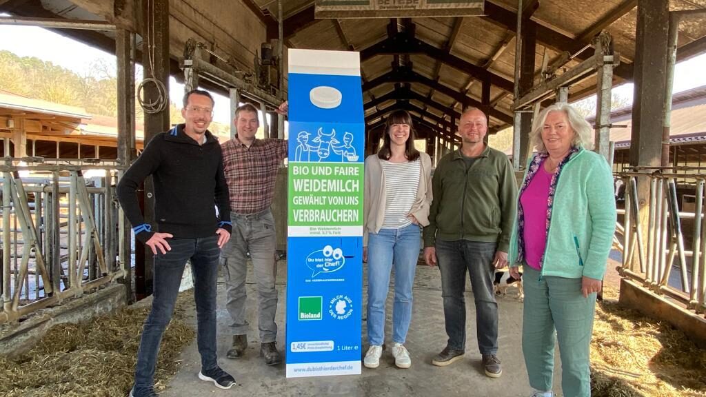 Menschen stehen im Kuhstall um eine große Packung "Du bist hier der Chef!"-Milch